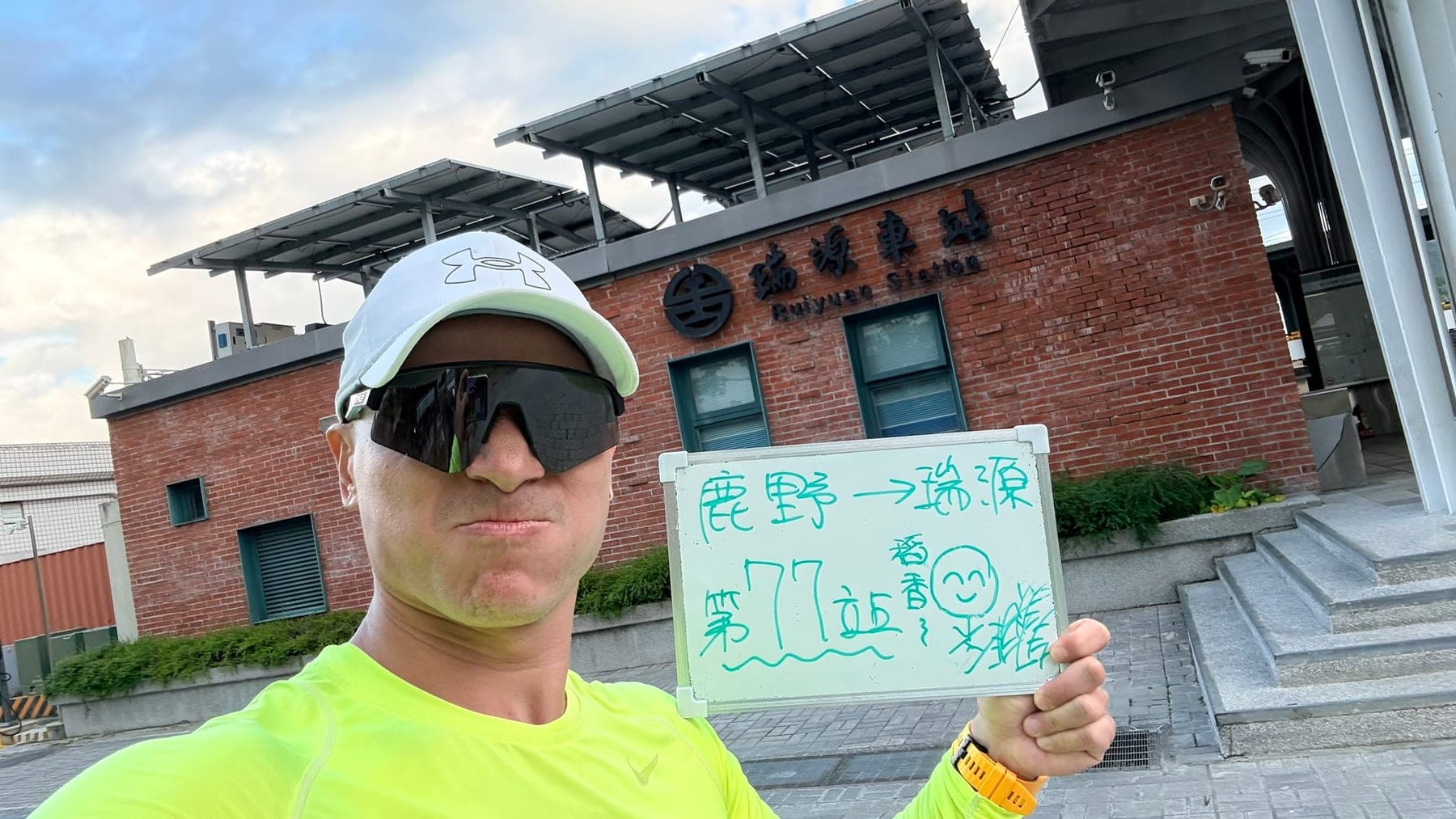 站主在瑞源站站牌前的走訪紀錄照，手持寫著「徒步環島 第 77 站　鹿野 → 瑞源」的白板