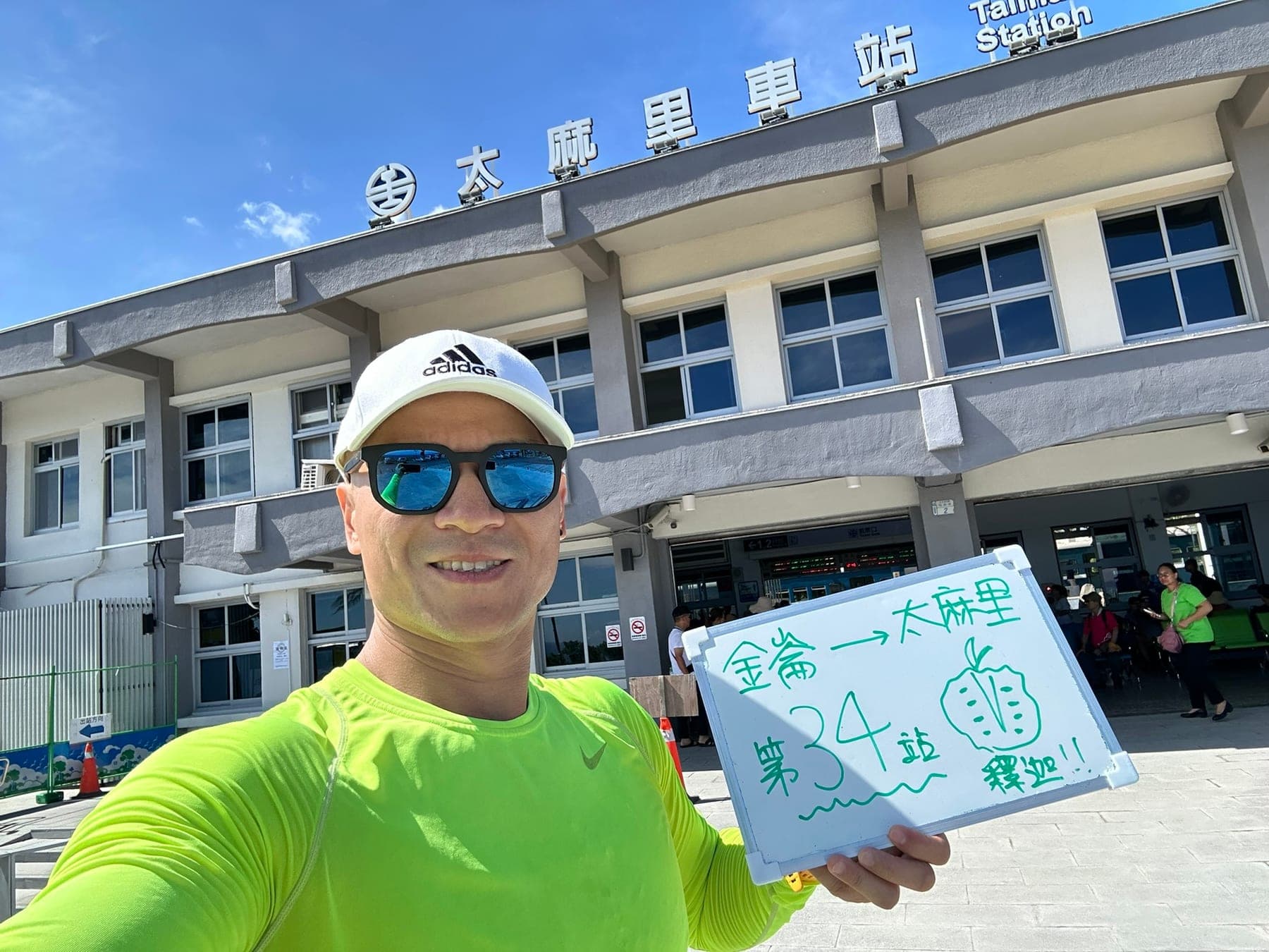 站主在太麻里站站牌前的走訪紀錄照，手持寫著「徒步環島 第 34 站　金崙 → 太麻里」的白板