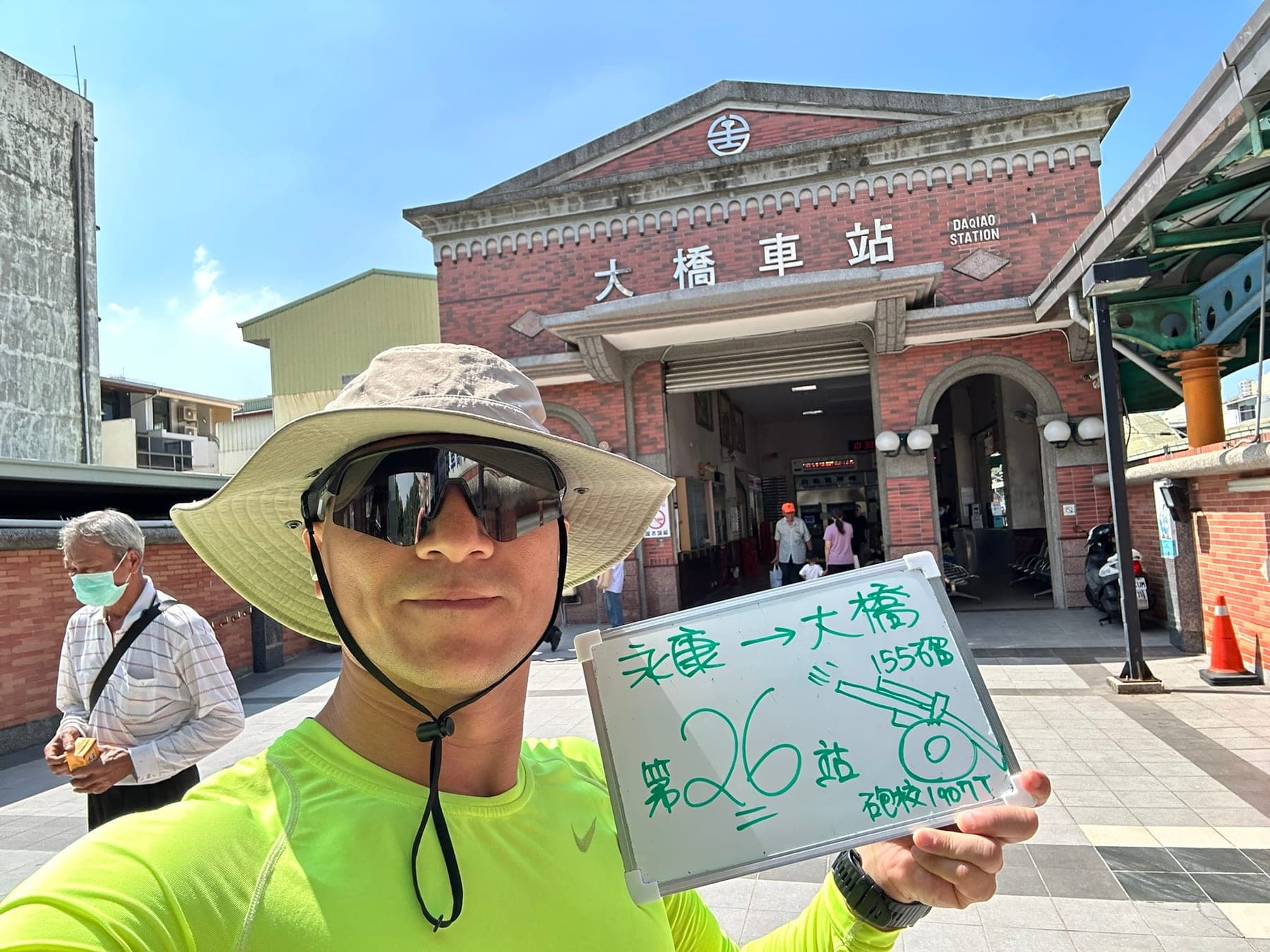 站主在大橋站站牌前的走訪紀錄照，手持寫著「徒步環島 第 26 站　永康 → 大橋」的白板