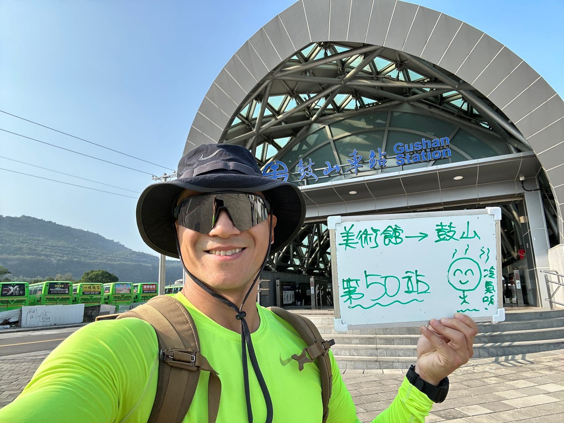 站主在鼓山站站牌前的走訪紀錄照，手持寫著「徒步環島 第 50 站　美術館 → 鼓山」的白板