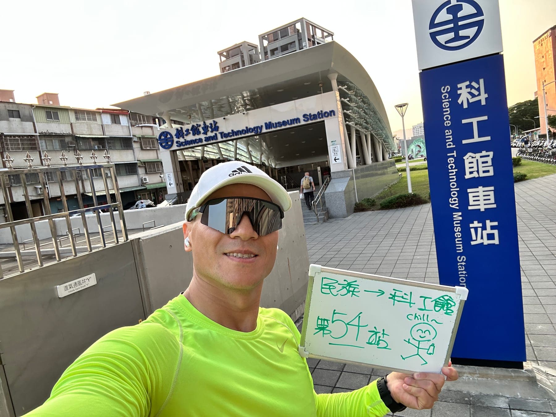 站主在科工館站站牌前的走訪紀錄照，手持寫著「徒步環島 第 54 站　民族 → 科工館」的白板