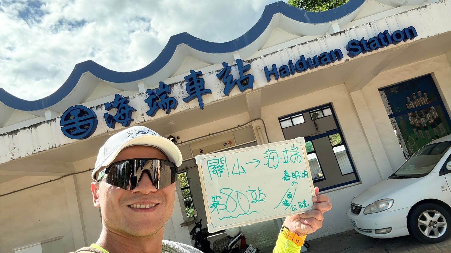 站主在海端站站牌前的走訪紀錄照,手持寫著「徒步環島 第 80 站 關山 → 海端」的白板