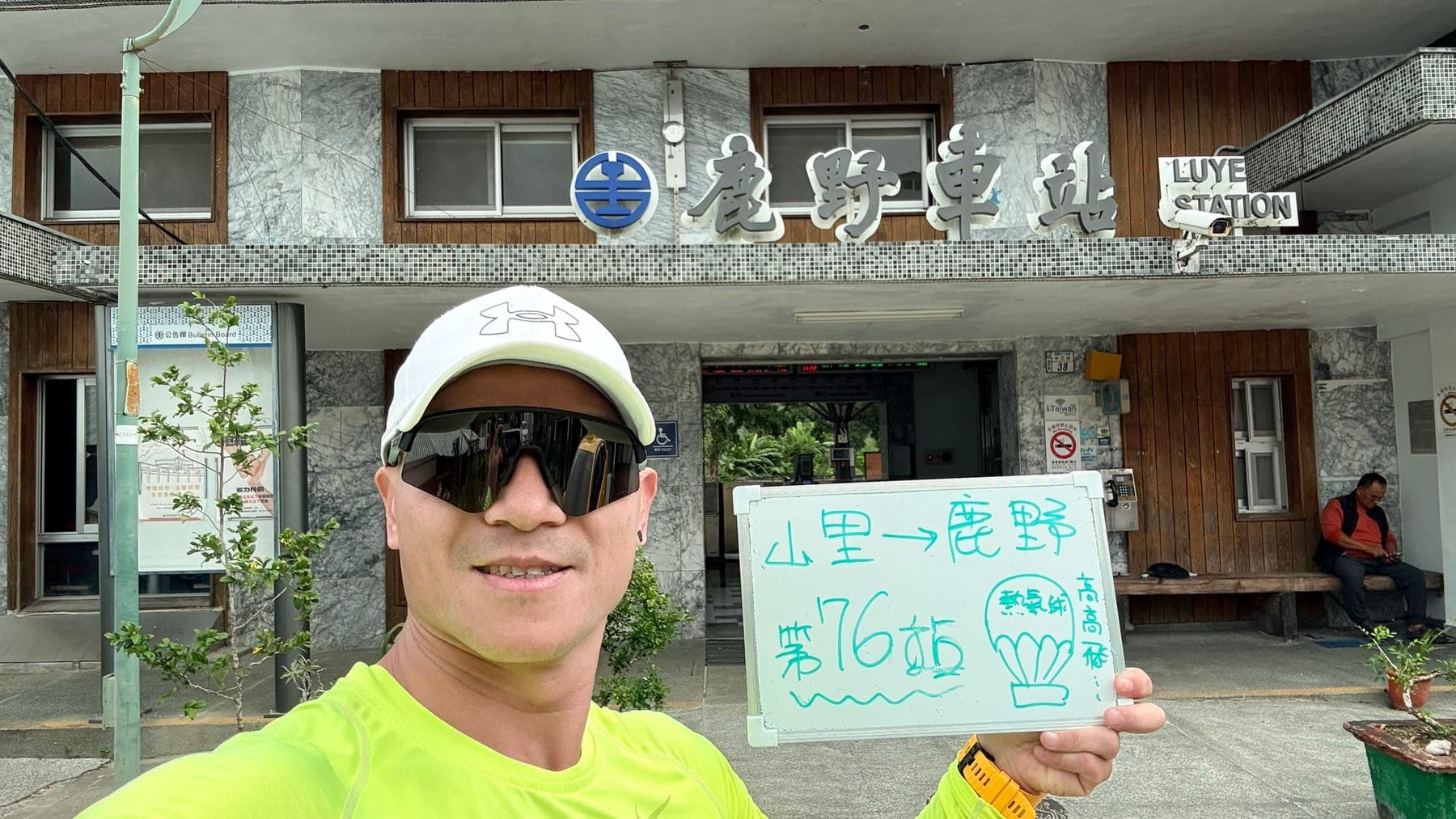 站主在鹿野站站牌前的走訪紀錄照,手持寫著「徒步環島 第 76 站 山里 → 鹿野」的白板