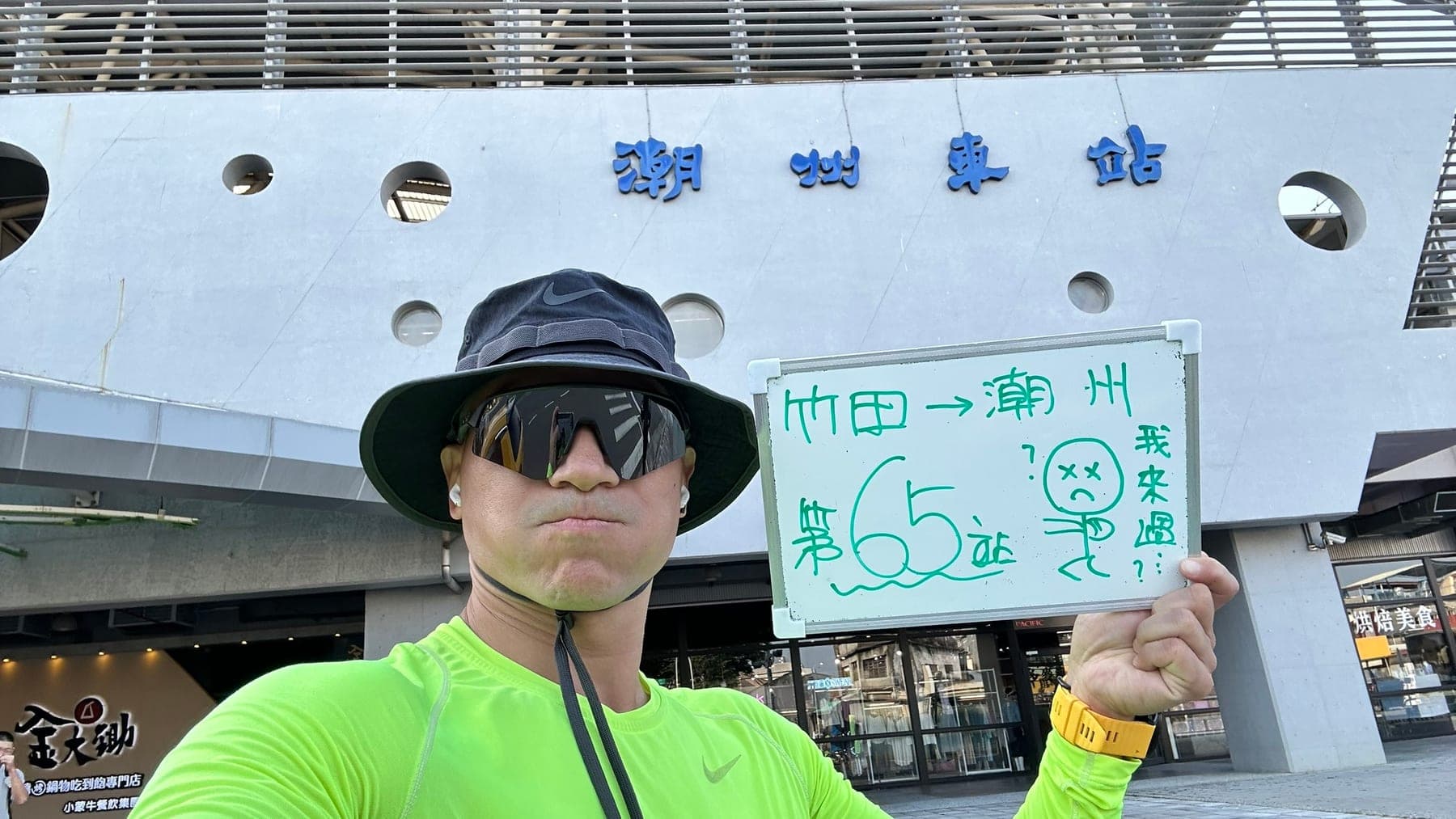 站主在潮州站站牌前的走訪紀錄照,手持寫著「徒步環島 第 65 站 竹田 → 潮州」的白板