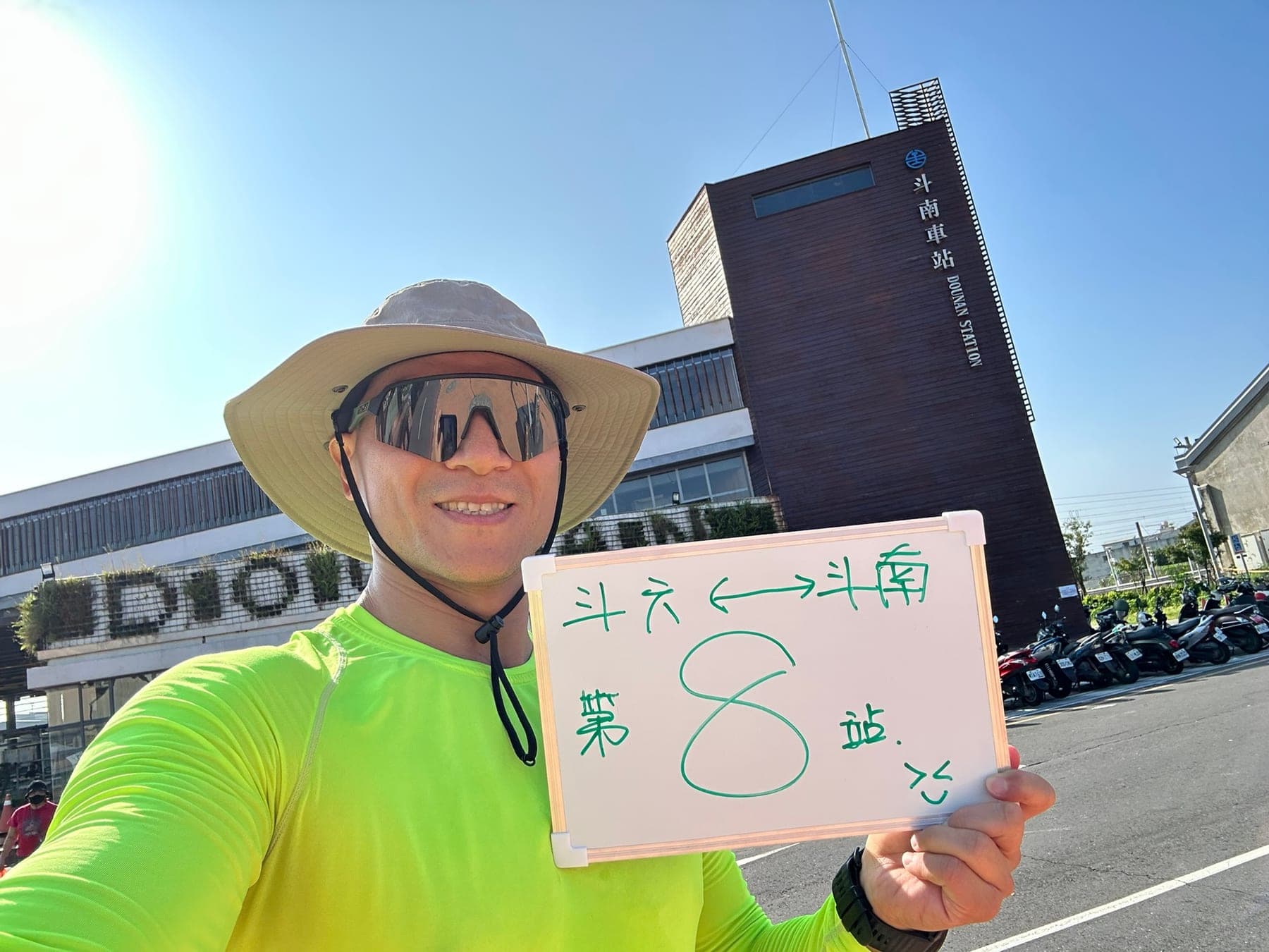 站主在斗南站站牌前的走訪紀錄照,手持寫著「徒步環島 第 8 站 斗六 ←→ 斗南」的白板