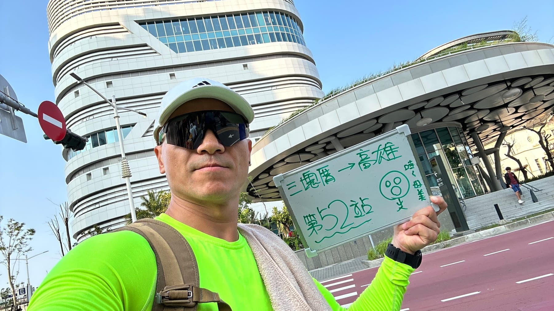 站主在高雄站站牌前的走訪紀錄照,手持寫著「徒步環島 第 52 站 三塊厝 → 高雄」的白板