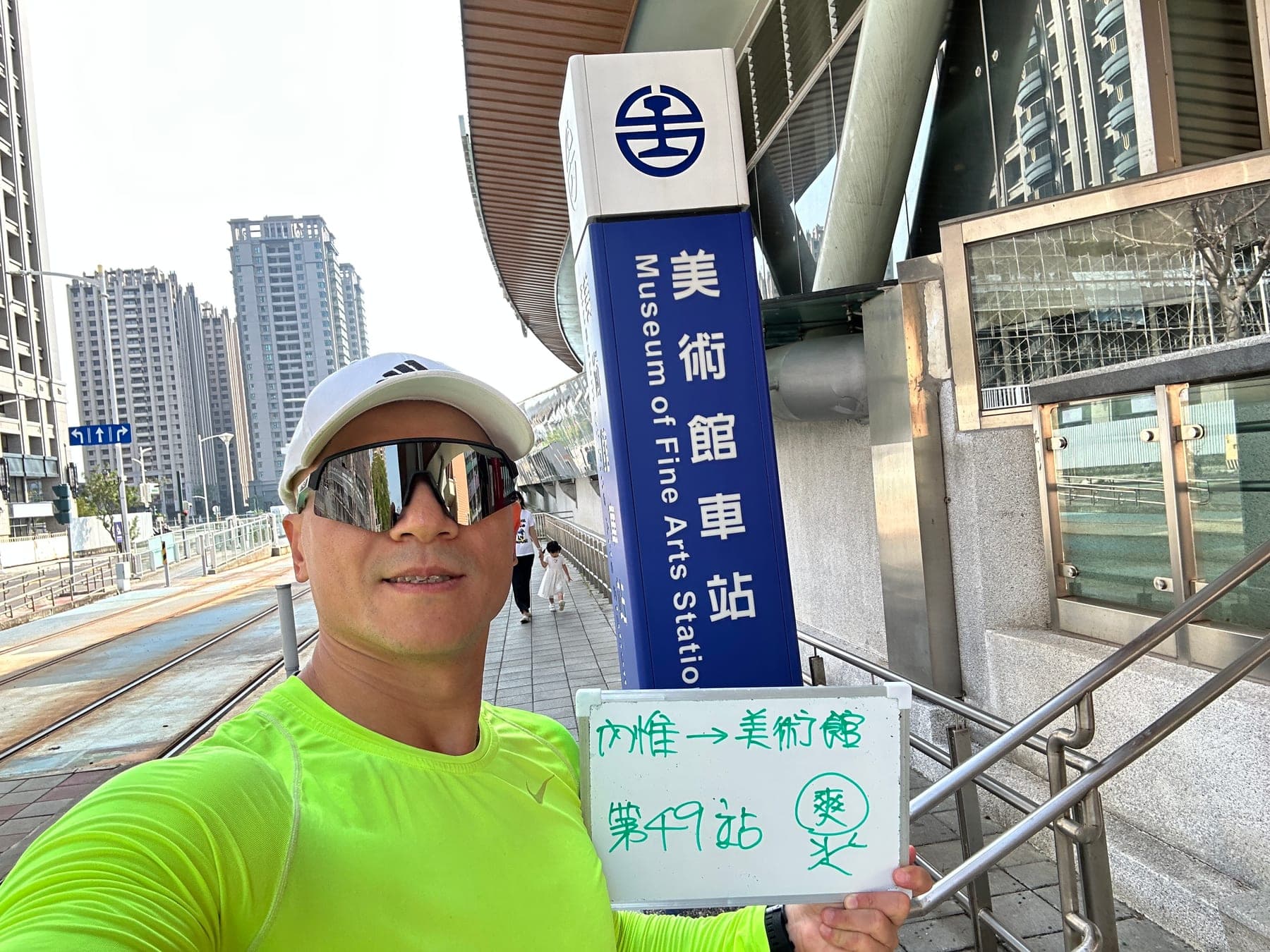 站主在美術館站站牌前的走訪紀錄照,手持寫著「徒步環島 第 49 站 內惟 → 美術館」的白板