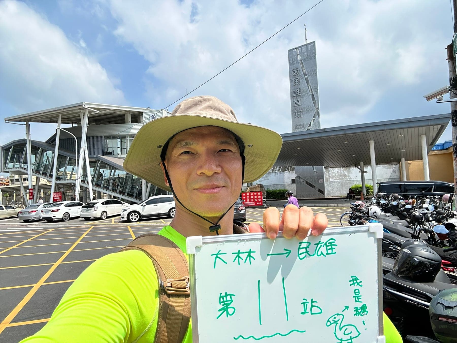 站主在民雄站站牌前的走訪紀錄照,手持寫著「徒步環島 第 11 站 大林 → 民雄」的白板