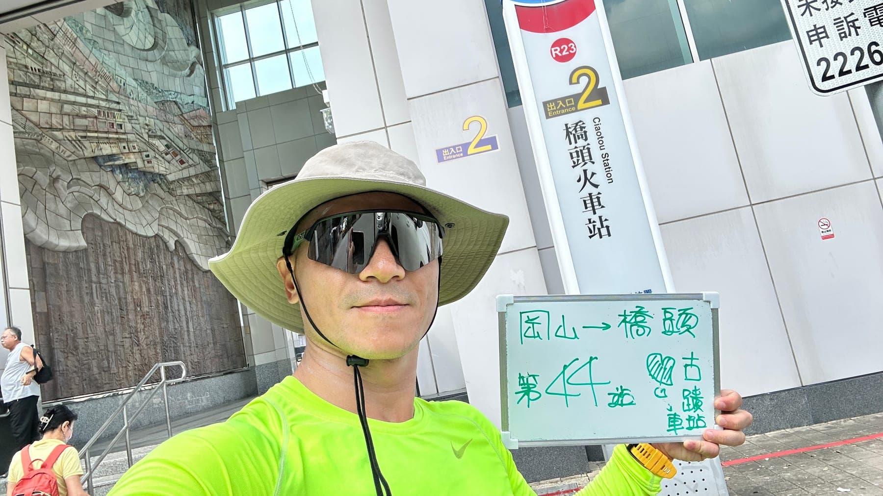站主在橋頭站站牌前的走訪紀錄照,手持寫著「徒步環島 第 44 站 岡山 → 橋頭」的白板