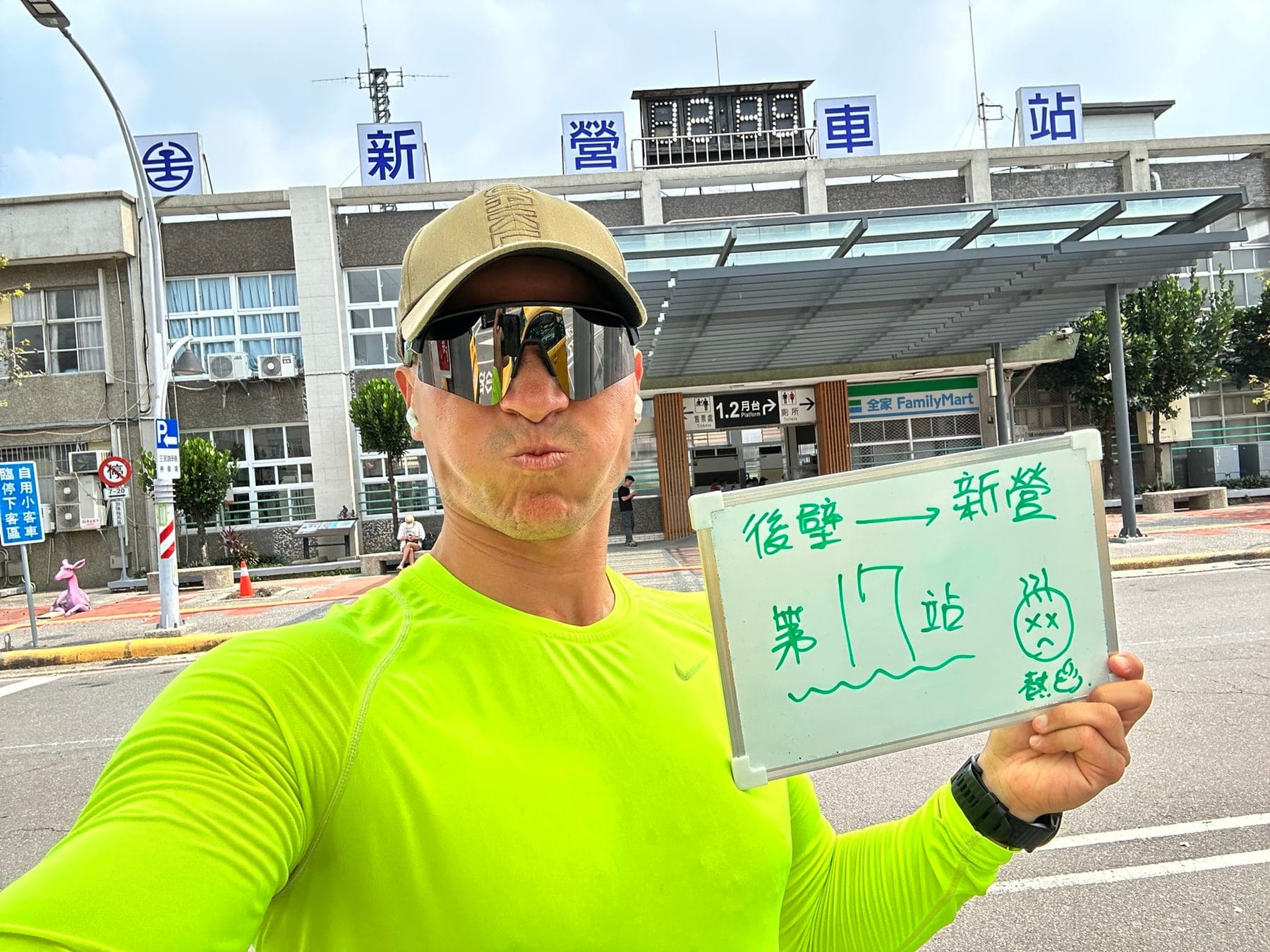 站主在新營站站牌前的走訪紀錄照,手持寫著「徒步環島 第 17 站 後壁 → 新營」的白板